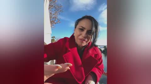 Video of a woman with straight black hair and fair skin wearing a red coat, standing outdoors with a clear blue sky and a tree in the background.