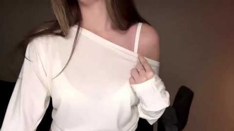 Video of a fair-skinned woman with long, light brown hair, wearing a white off-shoulder sweater, lifting the sleeve to reveal her shoulder, in a dimly lit room with a dark background.