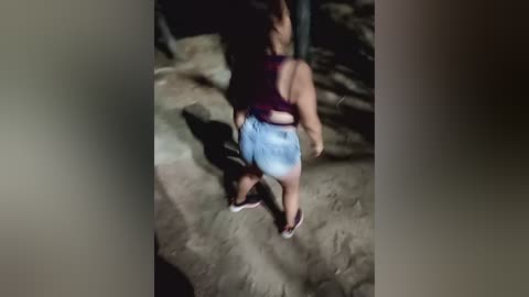 A video of a young woman with long, dark hair wearing a purple tank top and denim shorts, captured from behind, standing on a sandy, rocky surface.