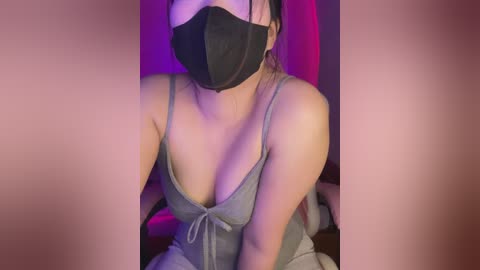 A video of a woman with light skin, wearing a black face mask and a low-cut gray tank top, sitting against a purple and pink backdrop.