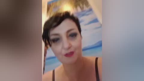 A blurry video of a fair-skinned woman with short, dark hair, wearing dark lipstick and a black spaghetti-strap top, standing in front of a beach-themed painting.