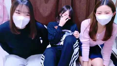 Video of three young East Asian women wearing white face masks, seated on a bed in a dimly lit room with brown curtains. They are dressed in casual attire, one in black, another in pink, and the third in navy.