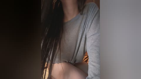 A video of a woman with long, dark braided hair, wearing a light grey crop top, partially visible, in a dimly lit room with a blurred background.