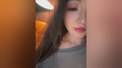 A video of an Asian woman with long black hair, light skin, and red lipstick, wearing a gray shirt. Background features warm lighting and a blurred interior with orange and brown tones.