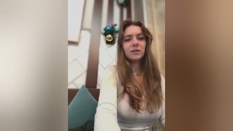 Media: Video of a young Caucasian woman with long, wavy blonde hair, wearing a white long-sleeve top, sitting in a modern, beige-toned room with geometric-patterned wallpaper and a teal chair.