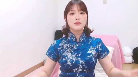 Video of an Asian woman with short brown hair, wearing a blue floral cheongsam, seated on a bed with pink sheets, in a minimalist bedroom with white walls.
