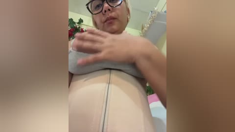 A video of a plus-size woman with short blonde hair, wearing glasses, lifting her grey bra to reveal her large breasts, standing in a brightly lit room with decorative plants.