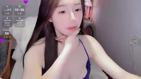 Video of a young East Asian woman with long black hair, wearing a blue tank top, sitting on a white chair. She touches her chin pensively, with a purple heart icon overlaid. Background shows a simple room with a white wall.