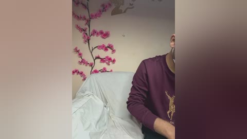 A video of a man with a beard, wearing a purple sweatshirt, sitting on a light blue couch. The background features a cherry blossom tree mural and a beige wall.