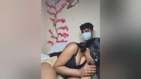 Video of a dark-skinned woman in a black top, wearing a mask, kissing a light-skinned man in a blue surgical mask, both sitting on a bed with pink cherry blossom wall art.