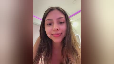 Media: Video of a young woman with long, wavy brown hair, light skin, and a slight smile, standing indoors with a white ceiling and purple accent lighting.