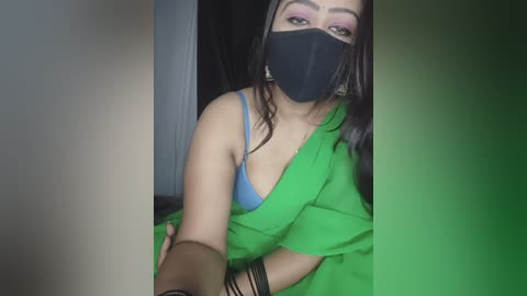 A video of a woman with medium skin tone, wearing a black face mask, green saree, and blue bra, sitting indoors, with a blurred background.