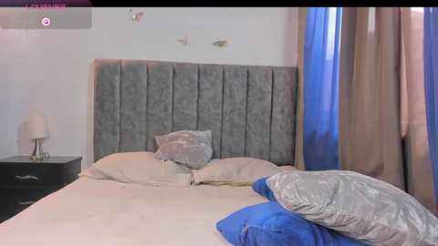 Video of a modern, minimalist bedroom with a grey, tufted headboard, beige bedspread, and blue pillows. Light-colored walls, a dark nightstand, and a lamp on the left side.