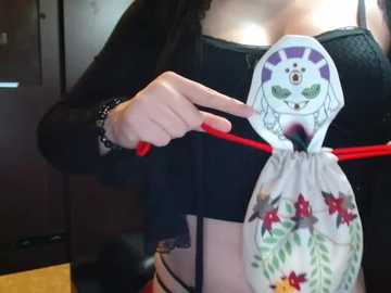 A video of a woman holding a decorative, embroidered handbag with a cartoon face, red flowers, and a black lace bra strap visible. She wears a black top.