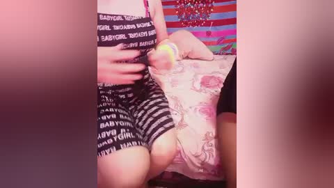 Video of a person wearing a black and white striped shirt, pink floral-patterned shorts, and pink panties, posing in a bedroom with colorful, patterned wall and bedspread.
