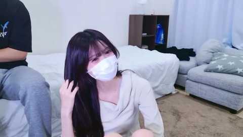 Video of a young Asian woman with long black hair, wearing a white face mask and beige sweater, sitting on a beige carpet in a living room with white furniture and a blue vase.