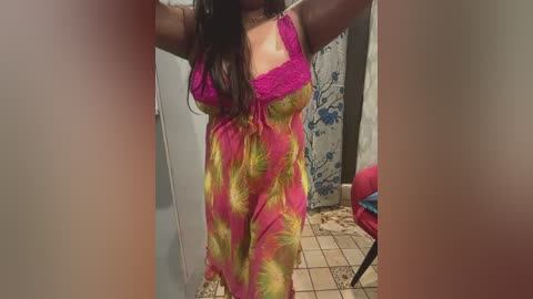 Video of a woman with long brown hair, wearing a vibrant pink and yellow tie-dye nightgown with lace trim, standing in a cluttered room with a tiled floor and a blue curtain.