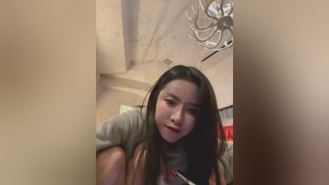 A video of an Asian woman with long black hair, wearing a grey t-shirt, standing in a dimly lit room with beige walls, an iron chandelier, and a red and white patterned wall.