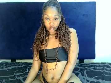 Video of a dark-skinned woman with long, curly brown hair, wearing a black strapless bra and black thong, seated on a patterned rug against a navy blue wall.