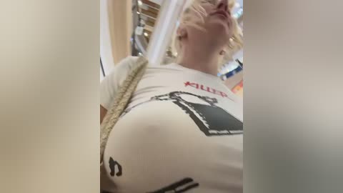 Video of a pregnant woman with blonde hair, wearing a white graphic T-shirt featuring a black and red design, walking indoors with blurred background.