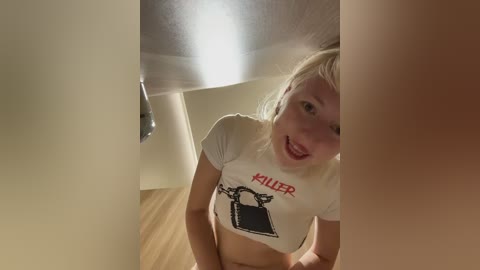 Video of a young blonde woman with a small frame, wearing a white \"Holler\" crop top, smiling inside a dimly lit hallway with wooden flooring.