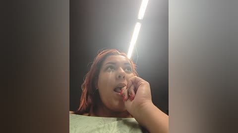Video of a young woman with red hair, light skin, and medium build, looking contemplative, resting her chin on her hand, under fluorescent lighting, against a dark background.