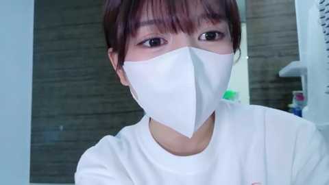 Video of an East Asian woman with short, dark hair, wearing a white surgical mask and plain white shirt, standing indoors against a gray wooden wall background.