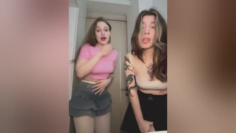 Video of two young women in a hallway, one with long brown hair, wearing a pink crop top and denim skirt, the other with short brown hair, a white tank top, and black skirt, both with surprised expressions.