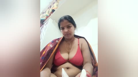 A video of a middle-aged Indian woman with medium skin tone, wearing a red bra and a patterned shawl, sitting on a bed, holding a white object, in a brightly lit room with a colorful curtain in the background.