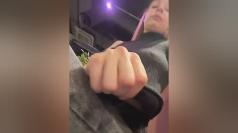 Video of a woman in a car, driving with a clenched fist on the steering wheel. She has short, pink hair and wears a gray sweater.