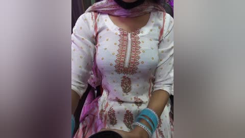 Video of a woman wearing a white embroidered kurta with pink and gold floral patterns, a matching pink dupatta, and blue bangles, seated indoors.