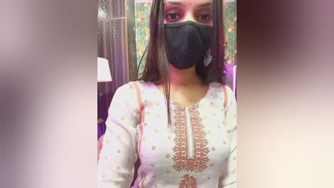 Video of a South Asian woman with long dark hair, wearing a black mask, white kurta with intricate embroidery, and dangling earrings, standing indoors with colorful floral wallpaper in the background.
