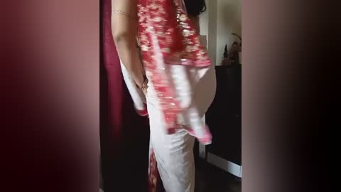 A video of a pregnant woman with a medium build, wearing a red sequined top and white pants, standing in a dimly lit room with a red curtain and a white door in the background.