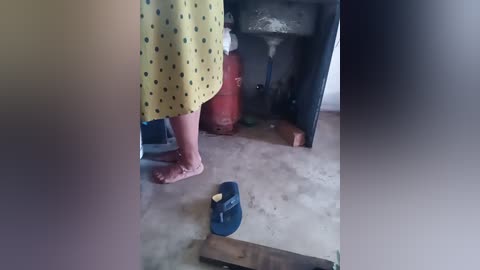 Video of a person wearing a yellow, polka-dotted dress, standing barefoot on a concrete floor. Nearby is a blue shoe, a red fire extinguisher, and a partially open wooden cabinet. The background includes a sink and pipes, suggesting a kitchen or utility area.