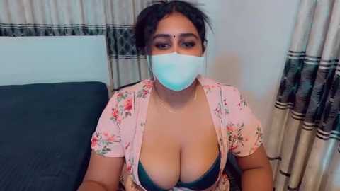 Video of a South Asian woman with medium-dark skin tone and dark hair in a floral blouse, revealing cleavage. She wears a blue bra, a surgical mask, and sits on a bed with white and beige curtains in the background.