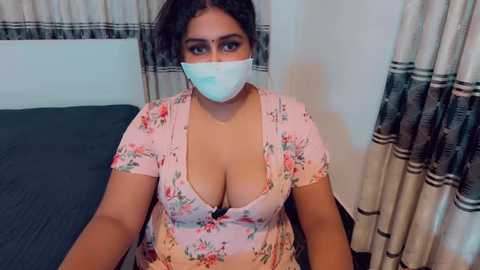 Video of a South Asian woman with dark hair, wearing a floral-patterned pink top with a deep V-neck, revealing cleavage, and a blue face mask. She sits on a dark bed in a bedroom with beige curtains.