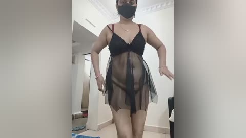 Video of a South Asian woman in sheer black lingerie, face mask, and red straps, standing in a dimly lit hallway with white walls and cream carpet.