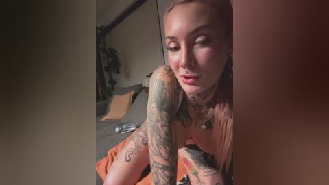 Video of a heavily tattooed, light-skinned woman with pink hair, wearing an orange top, sitting on a bed with orange pillows, in a dimly lit room.