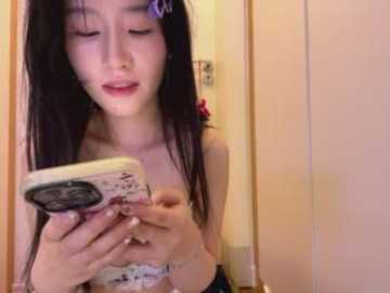 A video of an Asian woman with long black hair, wearing a white lace bra, taking a selfie with her smartphone in a dimly lit bathroom.