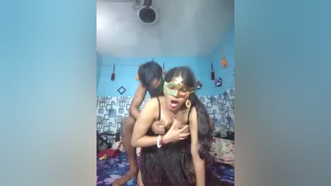 Video of a woman wearing a green mask and black dress, standing in a small, cluttered bedroom with blue walls and a patterned bedspread.