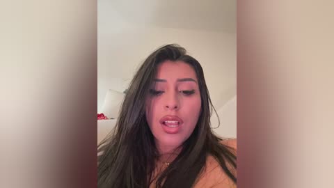 A video of a young woman with long, straight dark hair, wearing makeup, and a light-colored top, looking slightly to the left.