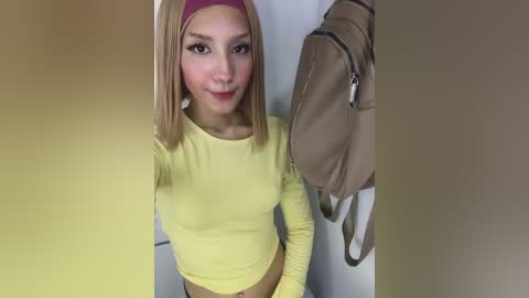 Video of a young, slim, light-skinned woman with straight blonde hair and a pink headband, wearing a tight yellow long-sleeved shirt and beige shorts, standing indoors, holding a tan backpack.