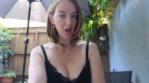 Video of a Caucasian woman with shoulder-length auburn hair, wearing a black lace bra, standing outdoors near a wooden fence and potted plants.