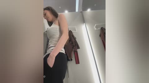 Video of a slender, light-skinned woman with straight brown hair in a white tank top and black sweatpants, standing in a modern, well-lit, minimalist restroom with white walls and recessed lighting.
