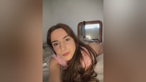 A video of a young woman with long brown hair and fair skin, wearing a pink top, lying on a bed in a dimly lit room, partially blurred.