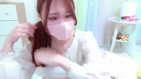 Media: A young woman with pale skin and long red hair, wearing a pink face mask and white knit sweater, sits on a white bed in a softly lit room with pastel curtains and colorful toys.