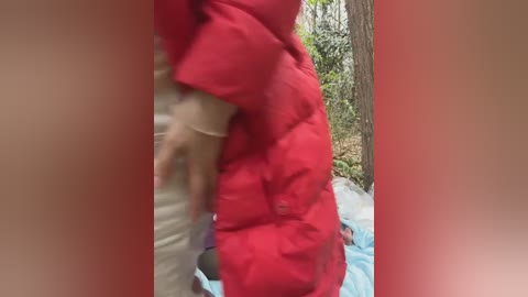 Video of a person in a bright red quilted jacket standing beside a light blue blanket in a forest setting with trees and foliage. The jacket has a puffy texture.