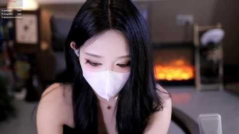 Video of an Asian woman with long black hair, wearing a white surgical mask, looking down, in a modern living room with a fireplace.