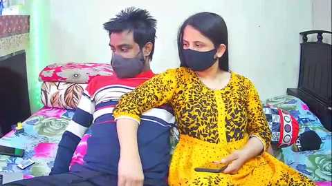 Video of a man and woman in face masks on a bed with floral-patterned sheets, wearing casual clothes. Background includes a wooden headboard and a bedside table.