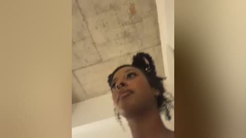 A video of a young Black woman with curly hair, wearing a nose ring, looking upward. She stands in a dimly lit room with beige walls and a concrete ceiling.
